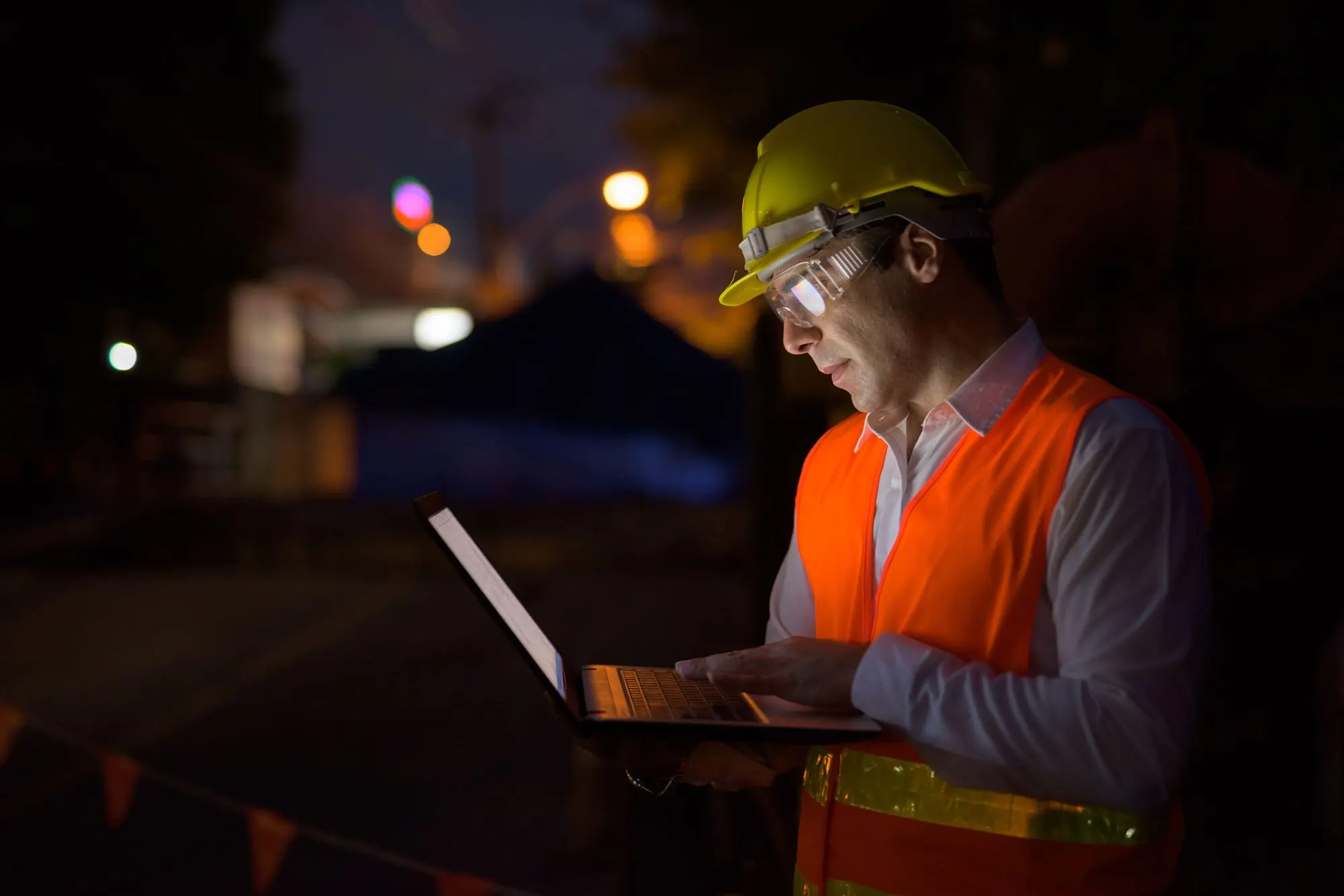 Engineer in a hard hat and high-visibility vest working on a laptop at night
